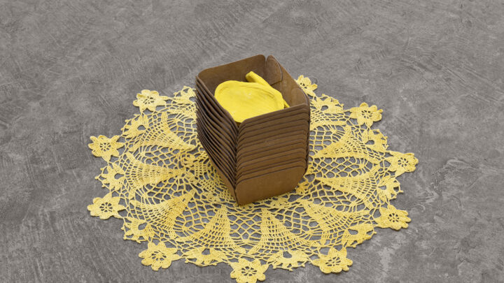 Sculptural work by Veronica Ryan titled 'Infection VII,' featuring a stack of brown paper containers with yellow objects inside, placed on a bright yellow lace doily on a gray surface.