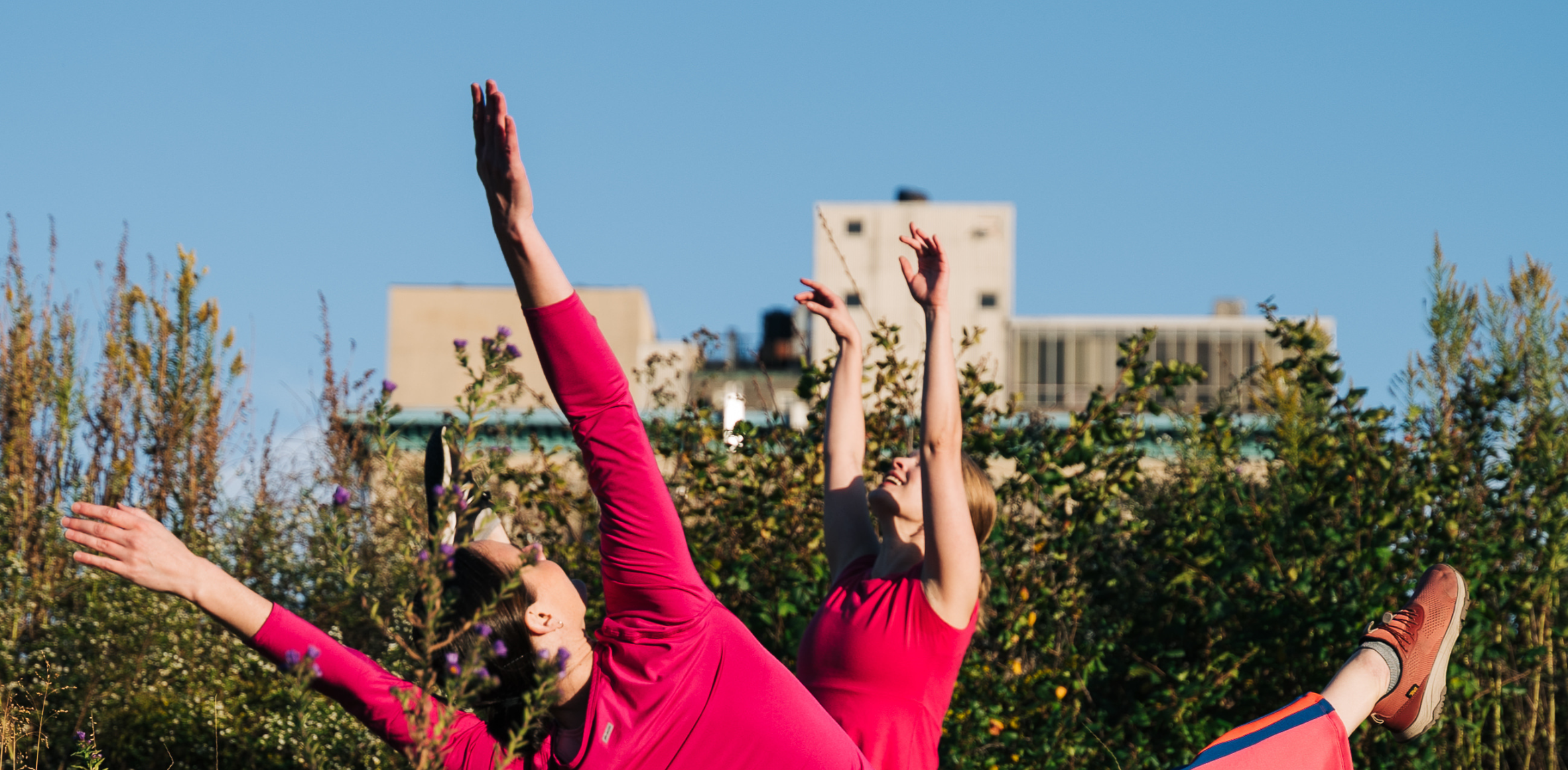 The image shows two individuals dressed in bright pink performing dance-like movements outdoors. Their arms are extended upwards, reaching towards the sky, and their bodies are tilted, suggesting dynamic motion. The scene is set in a lush, green environment, with tall plants in the foreground and urban buildings visible in the background, creating a contrast between nature and the city. The blue sky provides a clear, sunny backdrop.