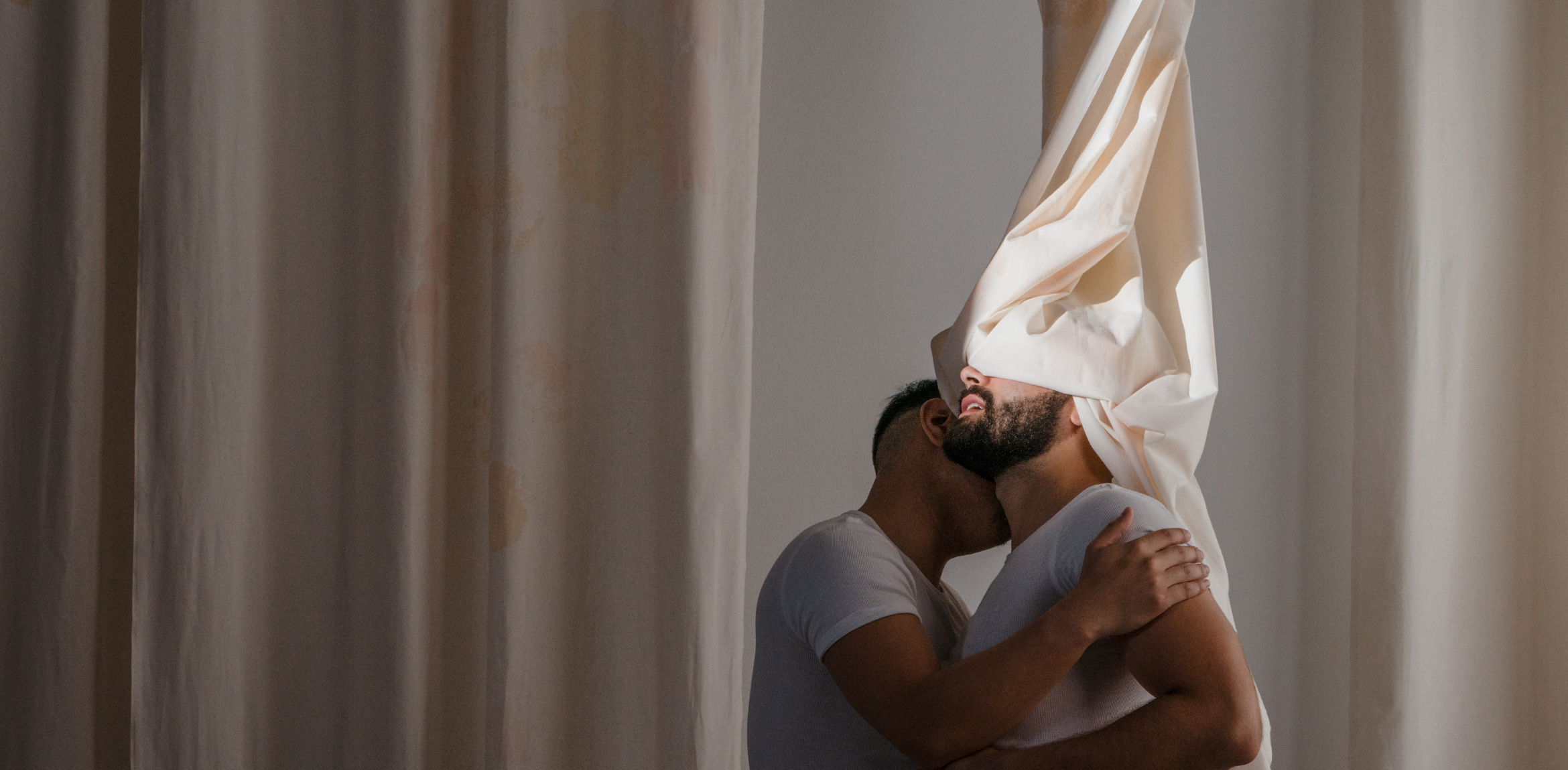 The image shows two individuals embracing intimately. One person, with a beard, has a large piece of fabric draped over their head, partially obscuring their face. The other individual, whose face is less visible, has their head close to the first person's neck. Both are wearing fitted white shirts. The scene is softly lit, with neutral-colored curtains in the background, creating a serene and private atmosphere. The fabric adds a flowing, dynamic element to the composition, enhancing the emotional and abstract feel of the moment.