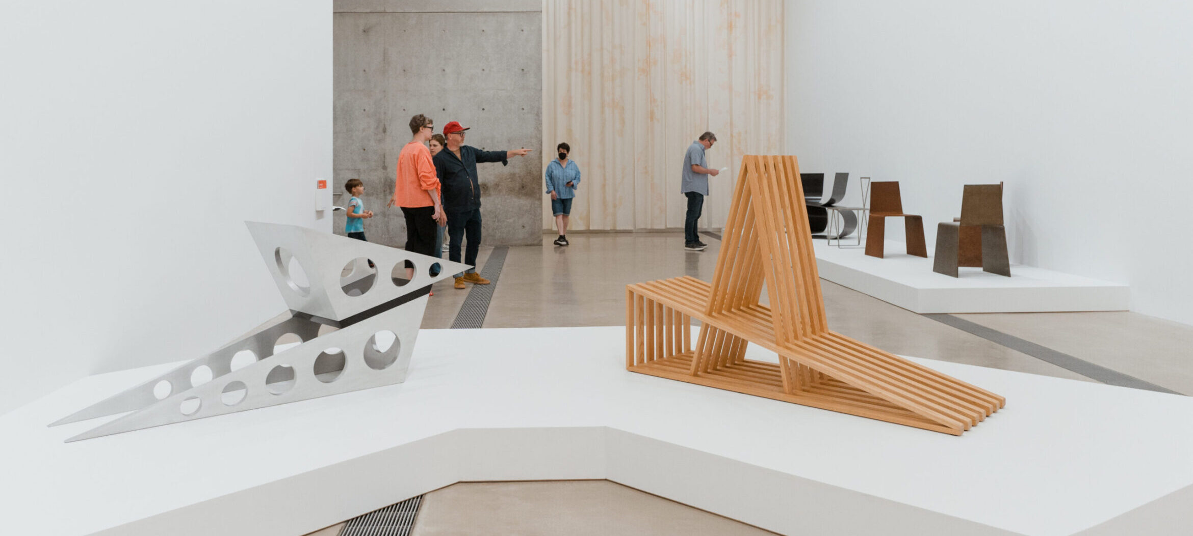 Two Adirondack inspired sculpture chairs on a white platform. There are people in the background observing additional furniture sculptures.