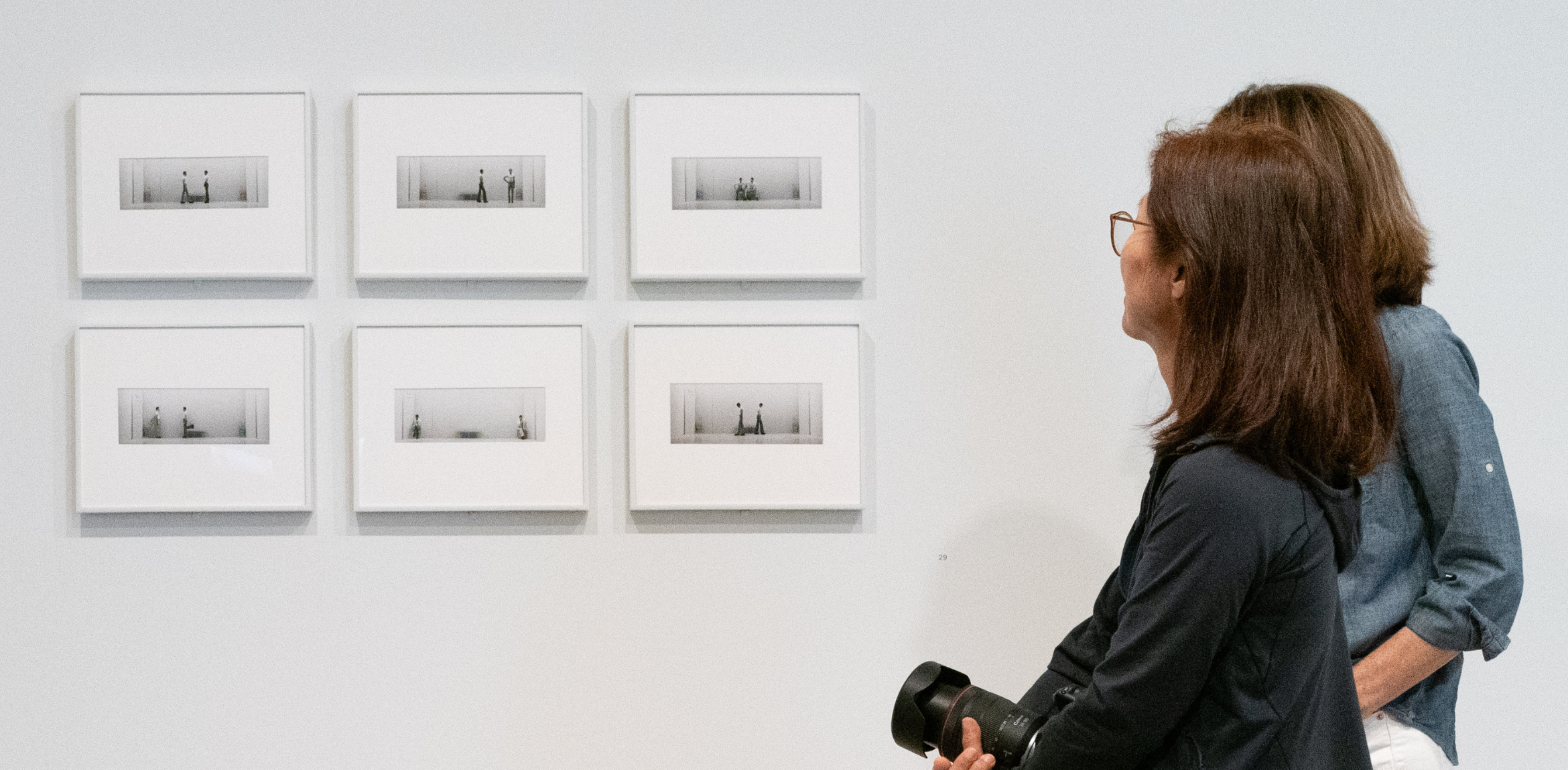 The image shows two women observing a display of six small, framed, rectangular black-and-white photographs arranged in two rows of three on a white wall. The women are standing side by side, with one holding a camera. The photographs appear minimalistic, possibly depicting figures in sparse environments. The overall atmosphere is one of quiet reflection in an art exhibition setting.