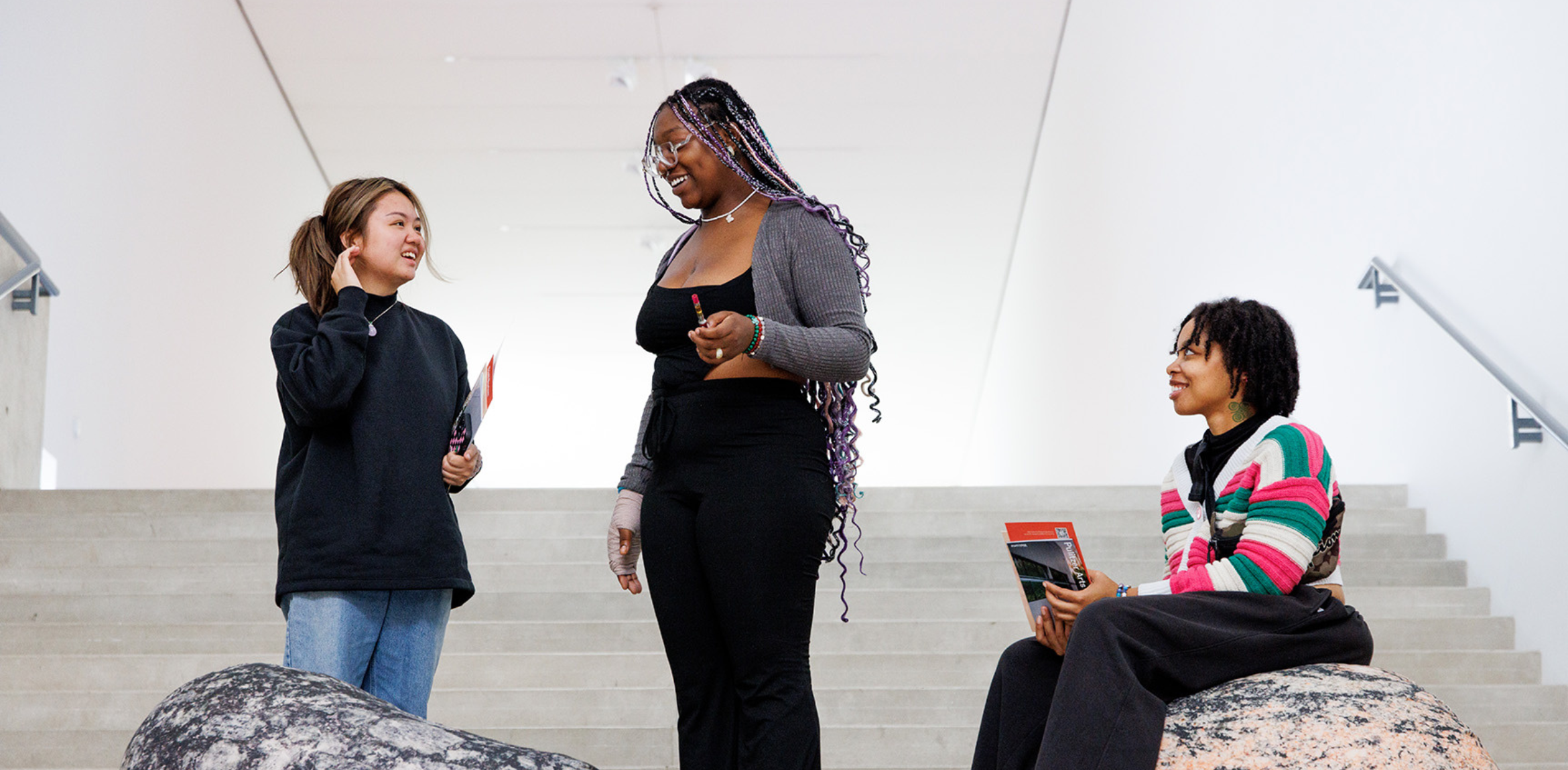 The image shows three young women interacting casually in what appears to be a modern, minimalistic space with wide stone stairs. One woman is sitting on a large textured stone, holding a brochure, while the other two stand nearby, engaged in conversation. The woman on the right is wearing a colorful striped sweater, while the others are dressed in more neutral tones. The atmosphere is relaxed and informal, suggesting a moment of friendly discussion, possibly at a cultural or academic event.