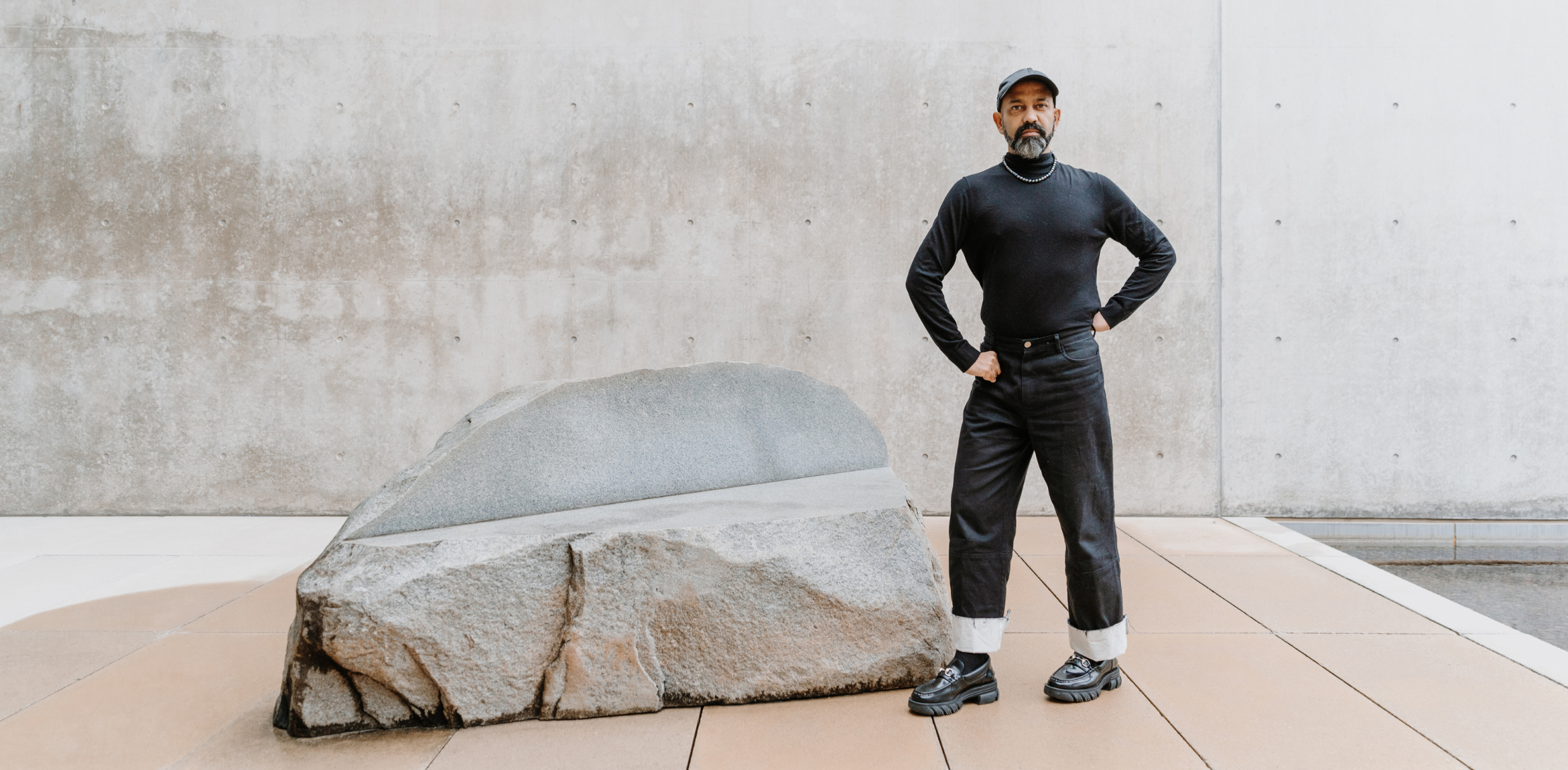 The image shows a person standing confidently with hands on their hips, wearing a black long-sleeved top, black pants with rolled-up cuffs, and chunky black shoes. The individual also has a beard and is wearing a black cap. Behind them, there is a large, smooth, flat-topped rock and a minimalist concrete wall, creating a modern, serene atmosphere. The overall setting appears to be an art or architectural space.