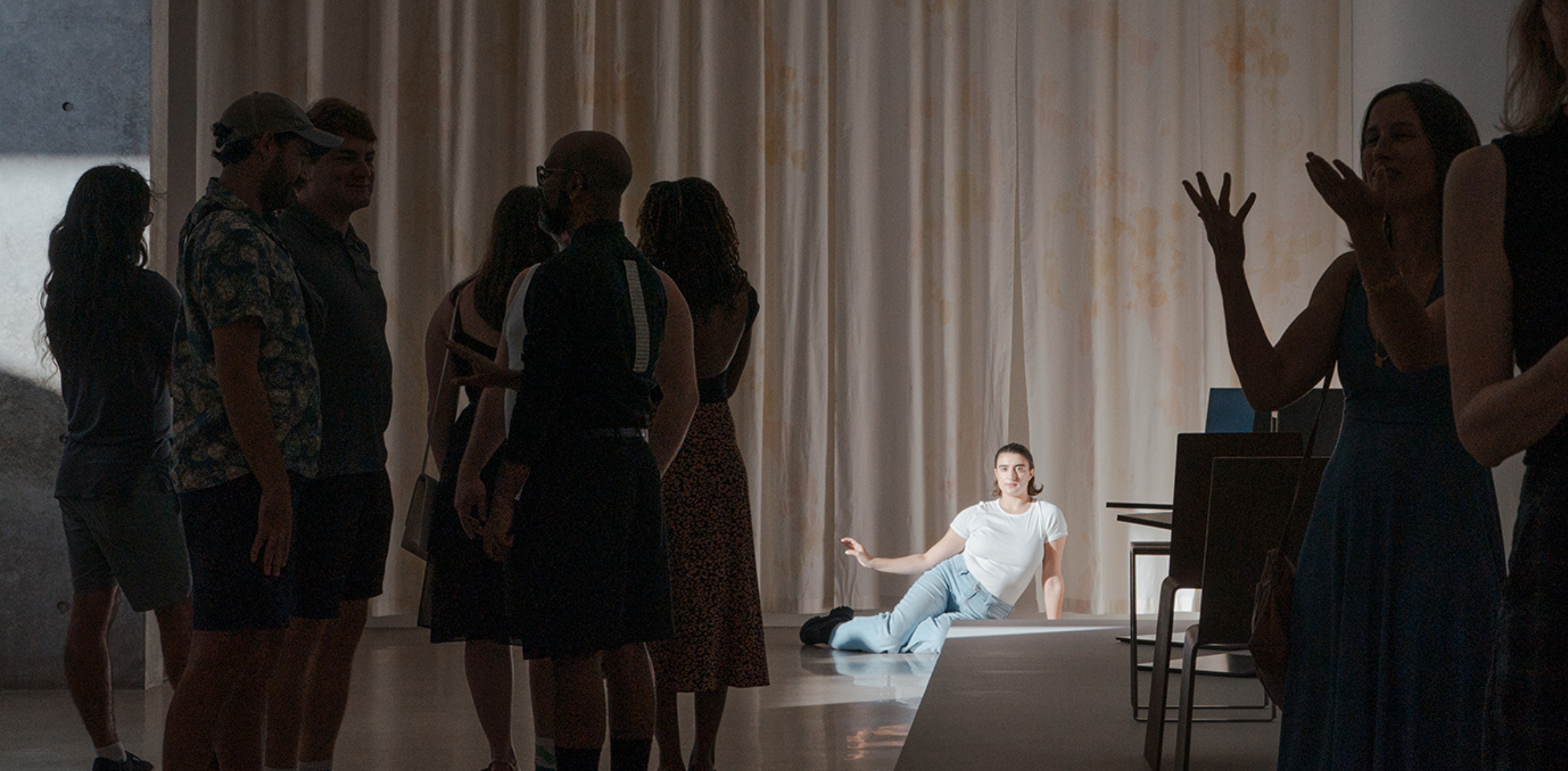 A performance scene with an audience in a gallery space. On the right, a performer dressed in a white shirt and light blue jeans sits on the floor, bathed in a spotlight. The audience, standing and talking amongst themselves, is gathered to the left. Some are in shadow, creating a contrast between the dimly lit group and the illuminated performer. The backdrop consists of light-colored drapes.