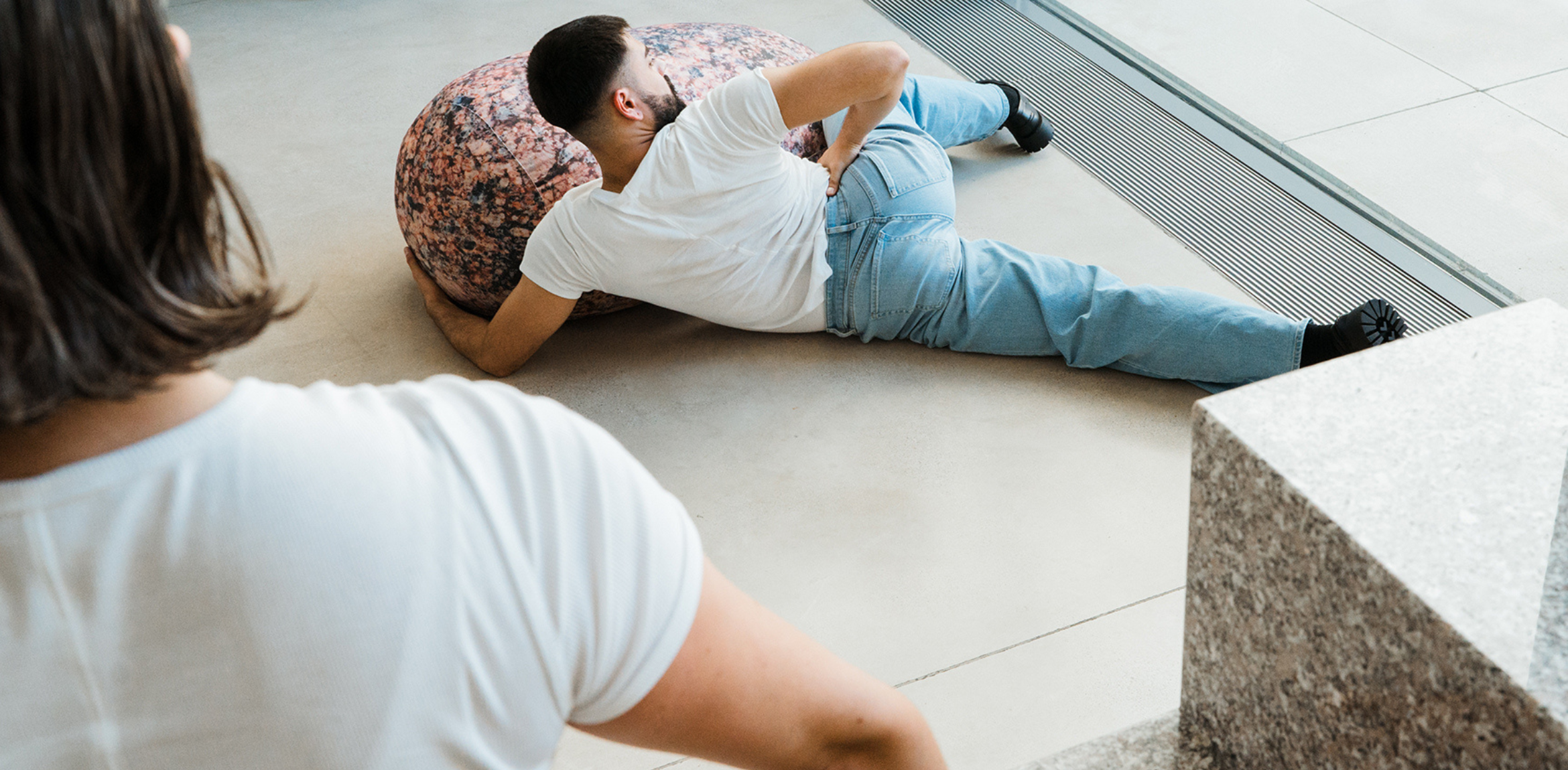 The image shows a person lying on the floor, leaning against a large, round, textured object resembling a rock. The individual is wearing a white T-shirt and light blue jeans. In the foreground, another person, also wearing a white T-shirt, is partially visible, observing or interacting with the scene. The setting appears to be indoors, near a window, with sleek, modern architectural elements visible. The image conveys a sense of movement or rest within a contemporary space.