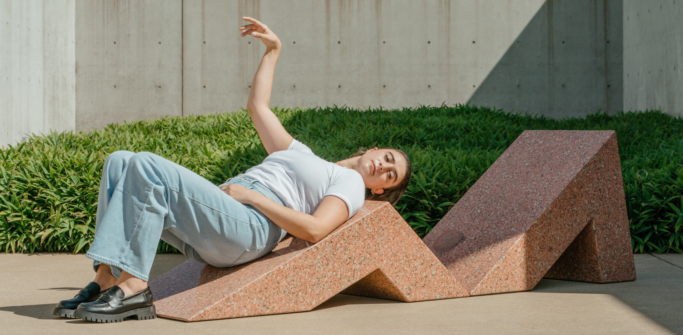 The image shows a woman reclining on a geometric, angular pink granite structure outdoors. She is dressed casually in light blue jeans and a white t-shirt, with black chunky loafers. Her body is positioned on the sculpture in a relaxed pose, with one arm raised above her head. In the background, there are lush green plants and a concrete wall, adding to the modern, minimalist setting. The scene gives off a casual, contemplative mood with a blend of human form interacting with architectural sculpture.