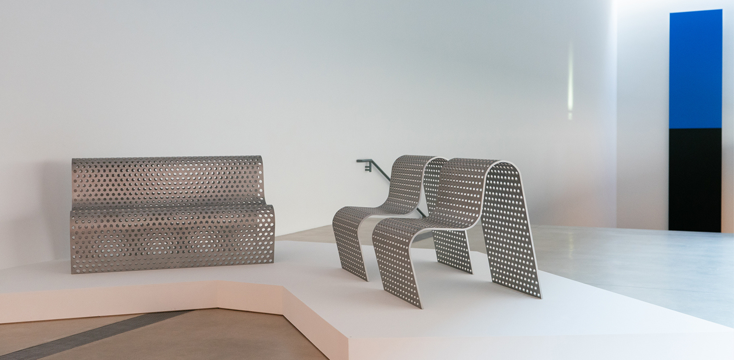 This image depicts modern, minimalist furniture composed of perforated metal sheets. The pieces include a bench and two chairs with sleek, curved designs. The furniture is displayed on a raised platform in a gallery-like setting with neutral tones, enhancing the industrial aesthetic of the metalwork. In the background, a geometric artwork featuring black and blue blocks adds contrast to the overall composition.