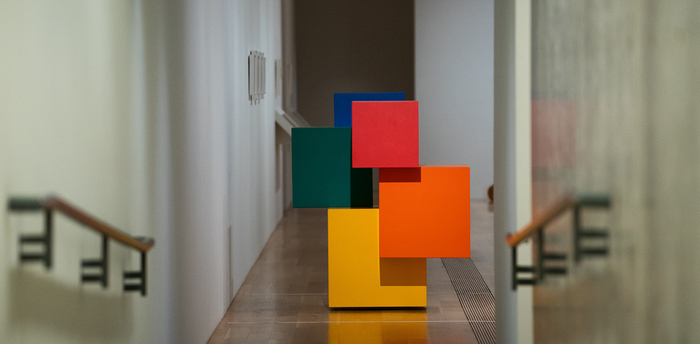 The image shows a geometric sculpture composed of brightly colored squares, stacked and arranged in a 3D form. The squares are colored green, blue, red, orange, and yellow. The artwork is displayed at the end of a hallway, with walls and handrails visible on both sides. The setting appears to be a gallery or museum, with natural lighting coming from above or the side. The overall mood of the space is calm and minimalistic, with the bold colors of the sculpture providing a stark contrast to the neutral surroundings.