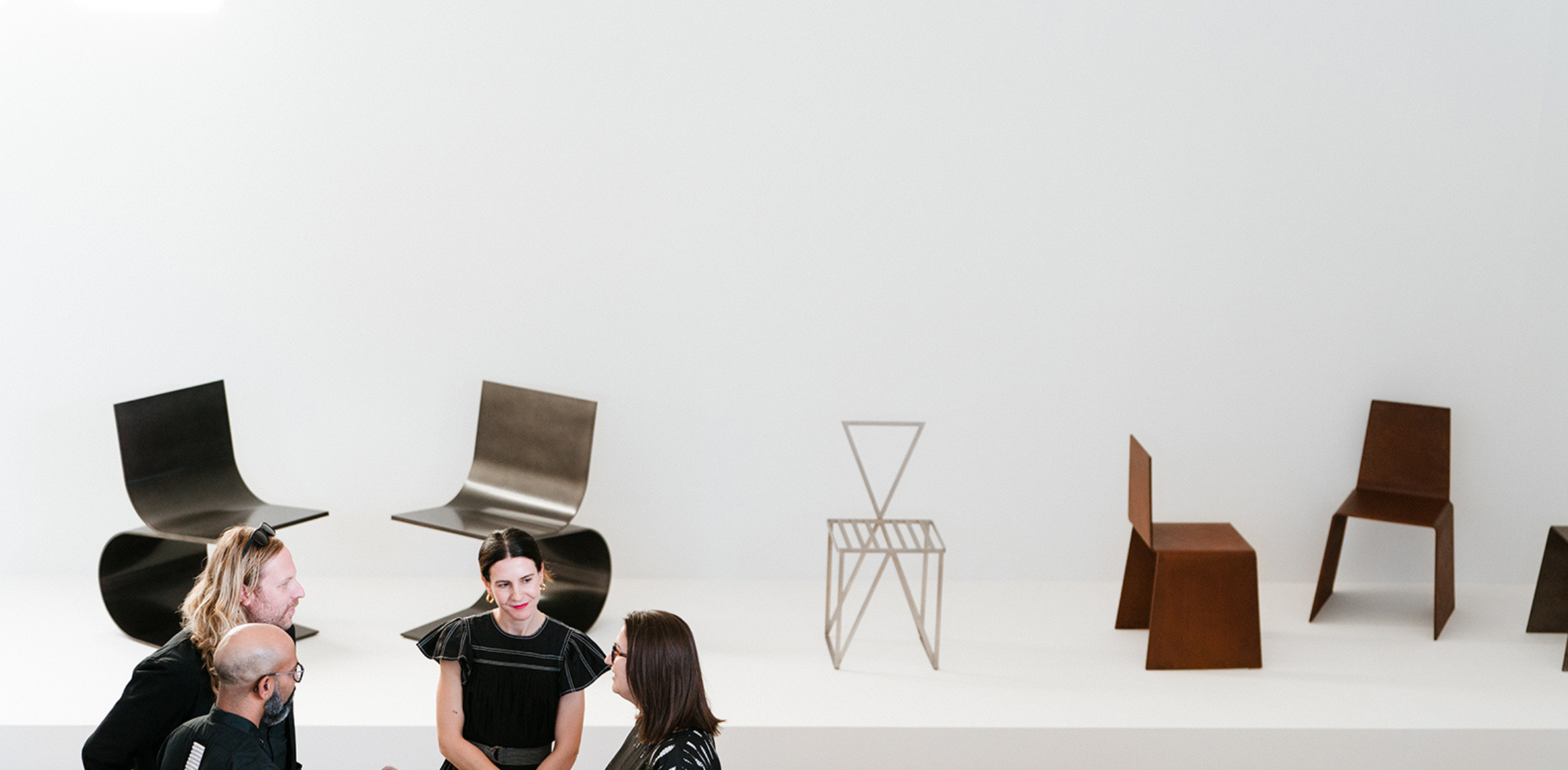 The image shows a gallery setting with a minimalist display of sculptural chairs. In the foreground, four people are engaged in conversation, while behind them, various modern chair designs, crafted from metal and wood, are showcased on a white platform against a clean, white wall. The overall feel is contemporary and artistic