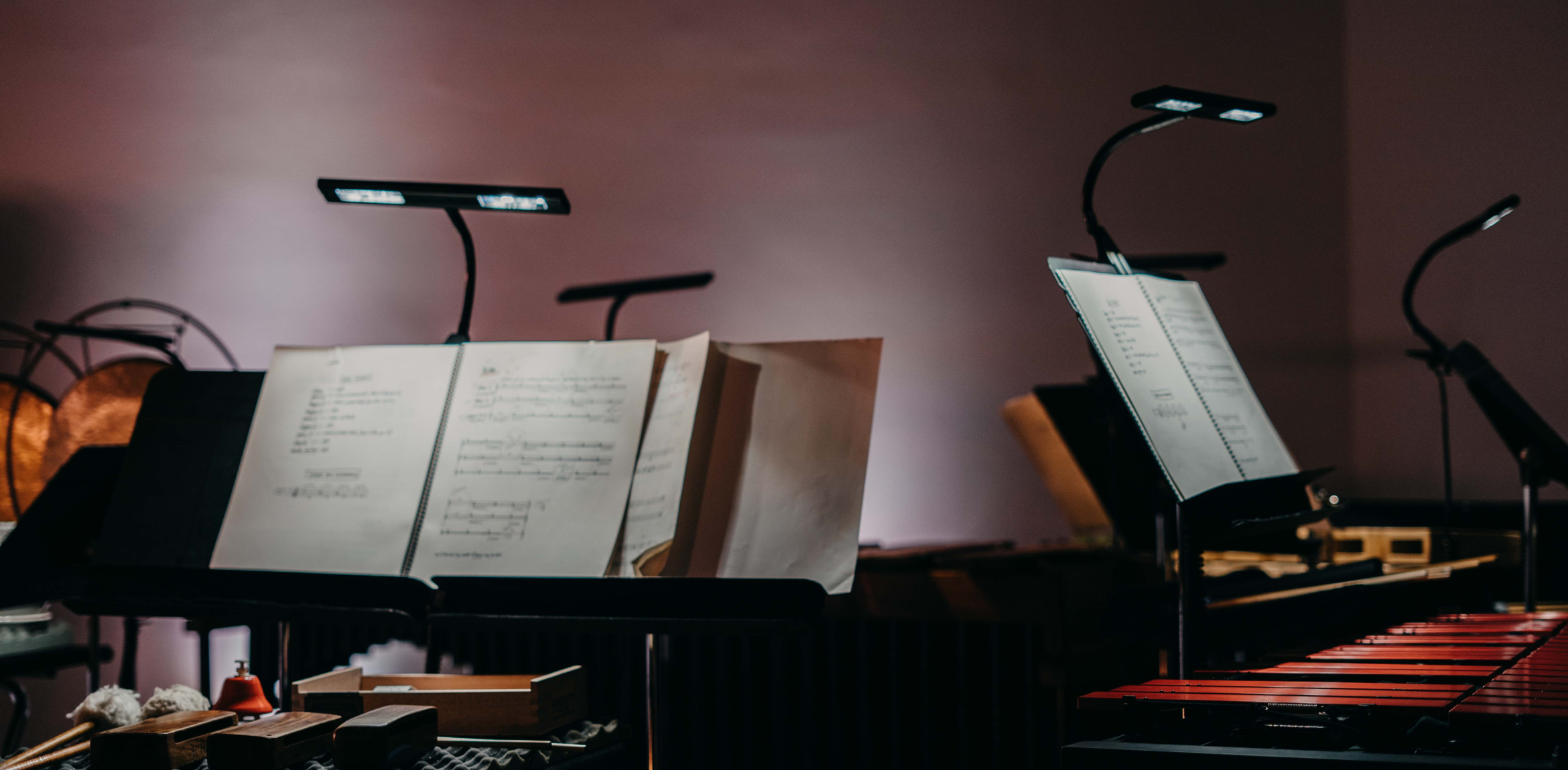 Three music stands hold open sheet music, illuminated by small, flexible lamps attached to each stand. The stands are black metal, likely adjustable in height. In the background, a gong and the red keys of a vibraphone are partially visible. The setting appears to be a dimly lit room, possibly a rehearsal space or stage.