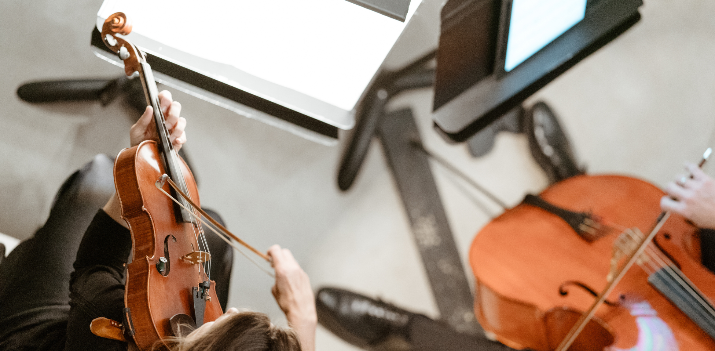 The image shows a close-up view of musicians playing string instruments, specifically a violin and a cello. The violinist's hands are visible as they hold the violin and bow, while the cellist's hand can be seen near the instrument's neck. Both musicians are seated, and music stands with sheet music are visible in the background, suggesting a rehearsal or performance setting. The lighting is soft, adding a warm tone to the image.