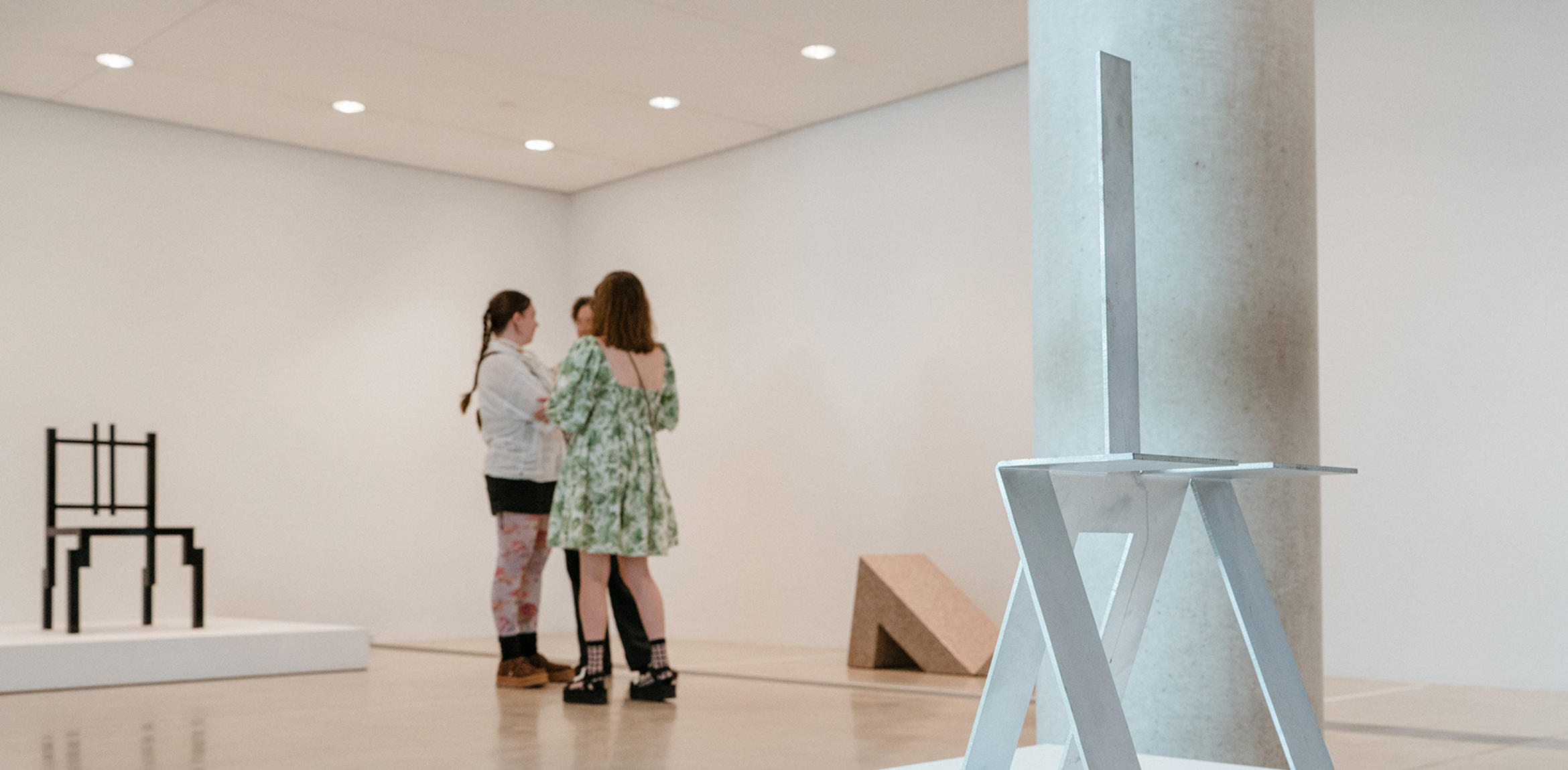 This image shows a modern gallery space where visitors are interacting. In the foreground, a minimalist metal sculpture with angular geometric lines is placed near a concrete pillar. In the background, two women are standing in conversation. Additional abstract sculptures can be seen on display, including a black rectangular structure and a triangular form in the distance. The white walls and simple lighting enhance the clean, contemporary feel of the exhibition.