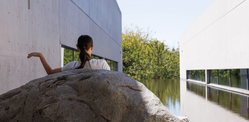 A person sitting on a large rock near the water courtyard of a modern concrete building, their arm gracefully extended to the side, framed by the reflection of trees and the minimalist architecture in the water