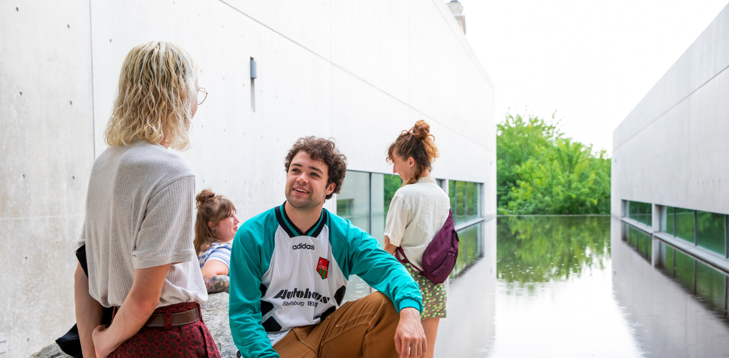 A group of four young people near a reflective pool between concrete buildings.