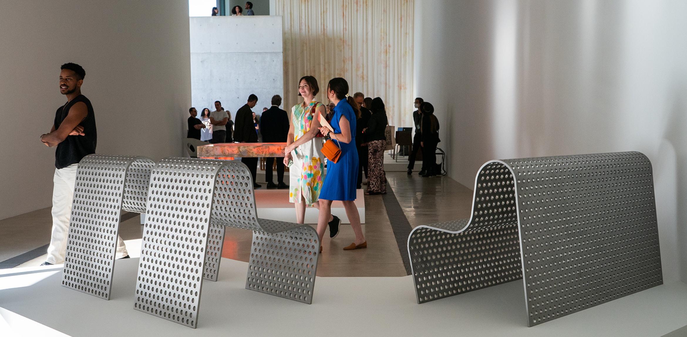 This image captures a lively scene at a gallery reception, featuring a perforated metallic sculpture in the foreground. Guests are seen mingling and admiring the artworks, with a softly lit, contemporary space in the background that includes additional sculptures and decorative elements. The atmosphere conveys an engaging and social art experience.