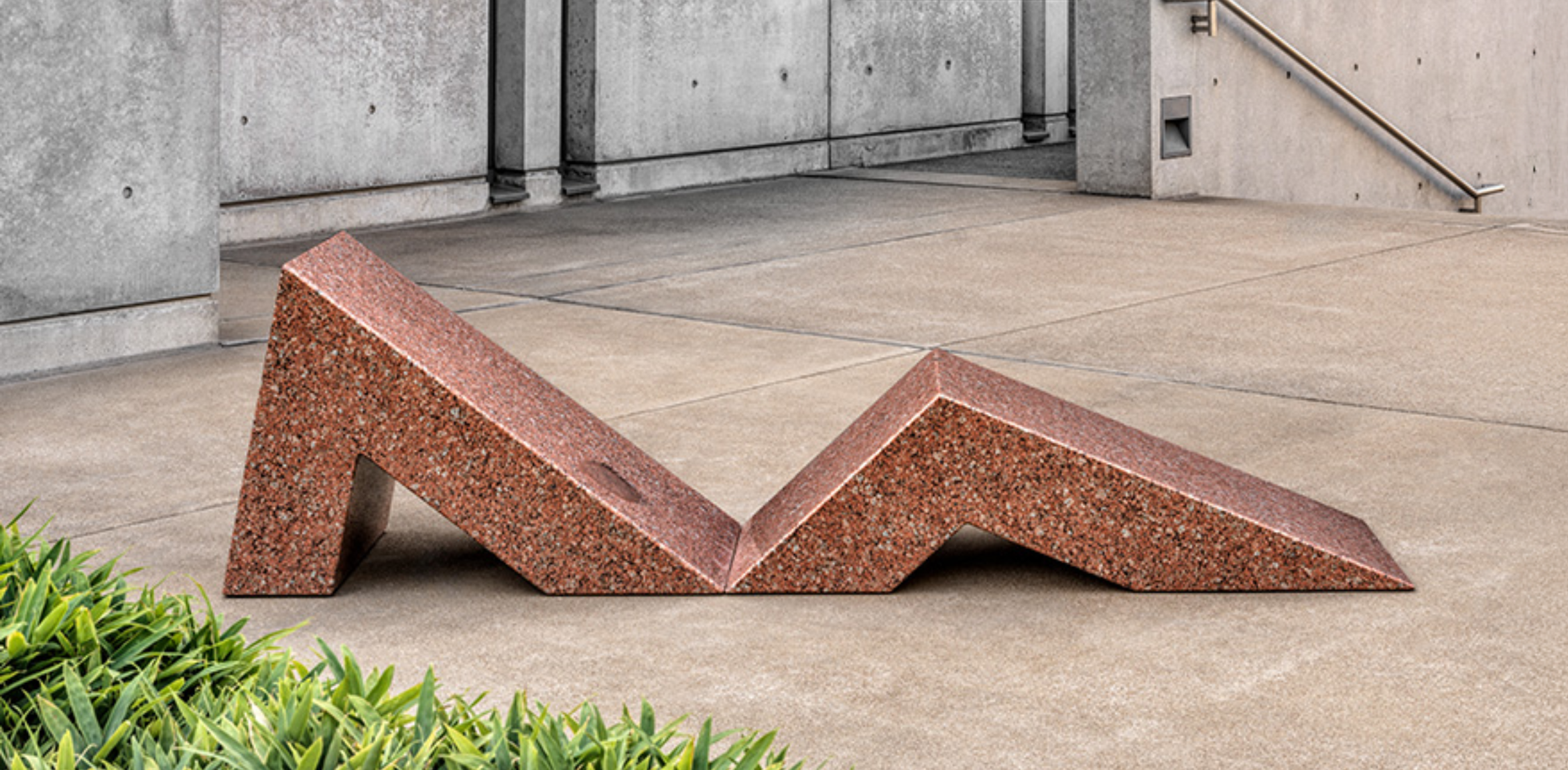 The image shows a geometric sculpture made of reddish-brown granite, placed on a paved outdoor surface near a building with concrete walls and a metal railing. The sculpture consists of angular, folded forms that create sharp peaks and valleys, contrasting with the greenery in the foreground. The setting is minimal and modern, emphasizing the material and design of the sculpture.