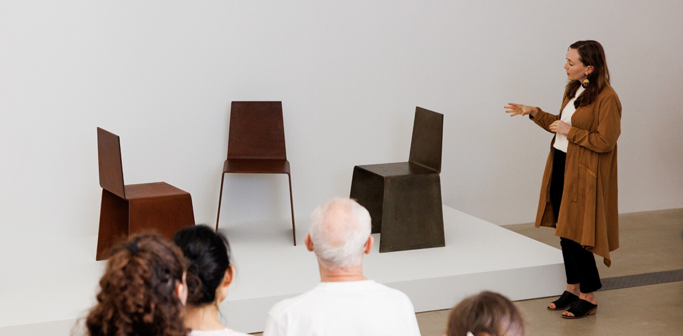 The image shows a woman giving a presentation to a small audience, standing beside a display of three minimalist chairs in a gallery setting. The chairs, made from metal, feature angular and geometric designs, placed on a white platform. The presenter, dressed in a brown long coat and black pants, gestures toward the chairs, engaging the seated viewers in front of her. The clean, neutral backdrop highlights the chairs as the focal point of the display.