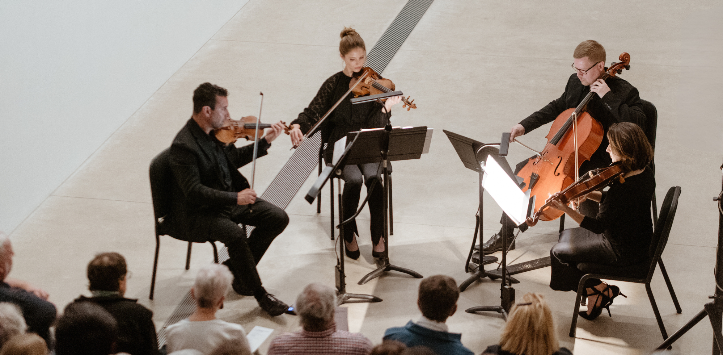 The image captures a quartet of musicians performing in an intimate setting. The ensemble, dressed in black, includes two violinists, a violist, and a cellist, all seated in a semi-circle. They are illuminated by music stand lights, creating a warm focal point against the minimal backdrop of a modern gallery space. The audience, partially visible in the foreground, attentively watches the performance, adding to the serene and engaging atmosphere.