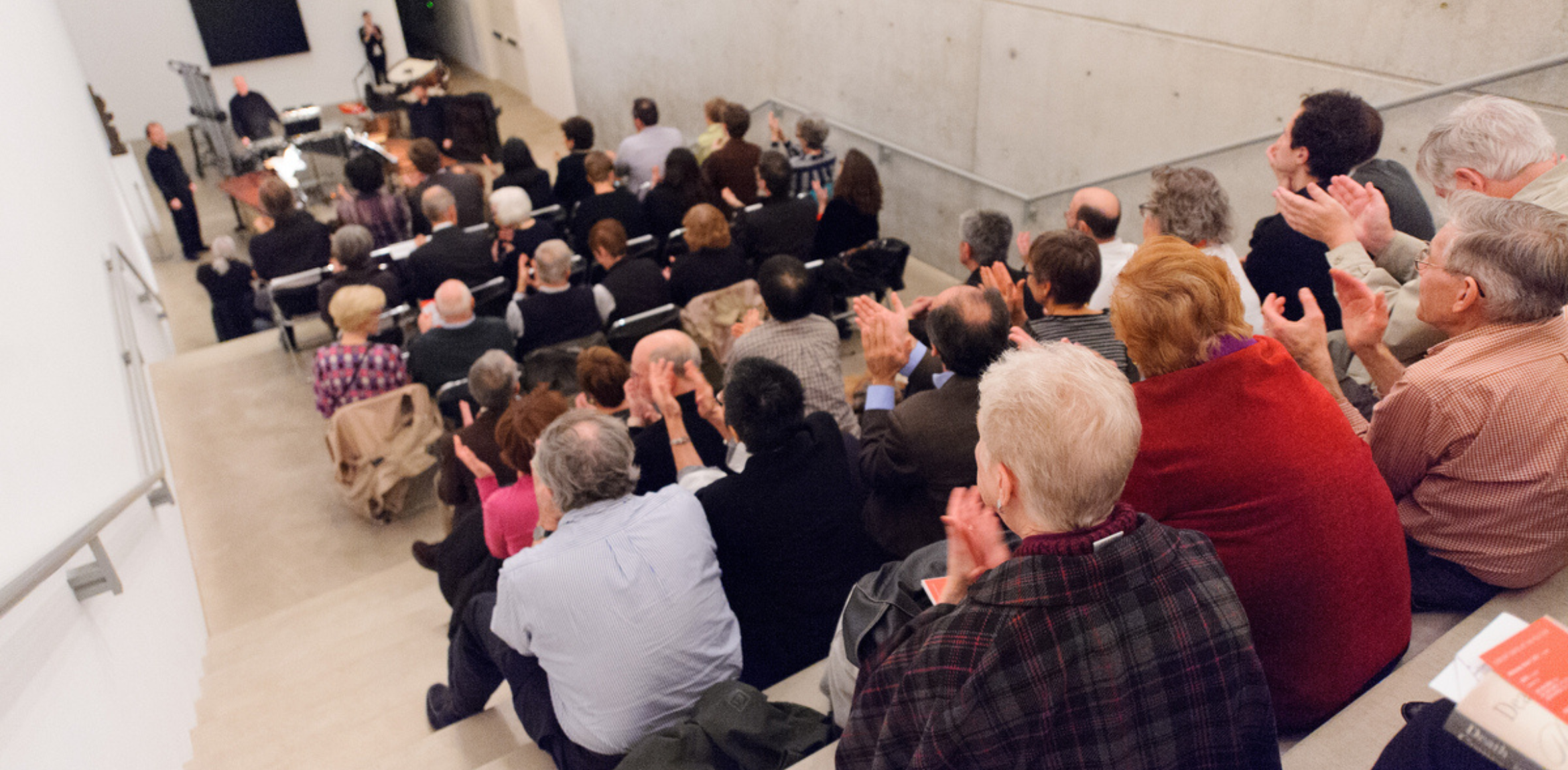 This image captures an audience seated in a tiered gallery space, applauding a live performance. The setting features a modern, minimalist interior with concrete walls and a focus on the engaged crowd, creating a lively and communal atmosphere.