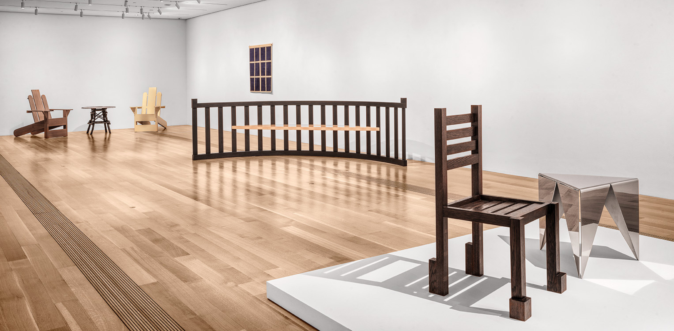 The image showcases an art gallery installation featuring a variety of sculptural furniture pieces arranged across a hardwood floor. In the foreground, a wooden chair with geometric detailing and a metallic angular stool are displayed on a white platform. Toward the center, a curved wooden bench and two uniquely designed chairs, one natural wood and the other painted yellow, are arranged alongside a small round table with intricate legs. The clean, minimalistic backdrop and strategic lighting highlight the craftsmanship and design of each piece.