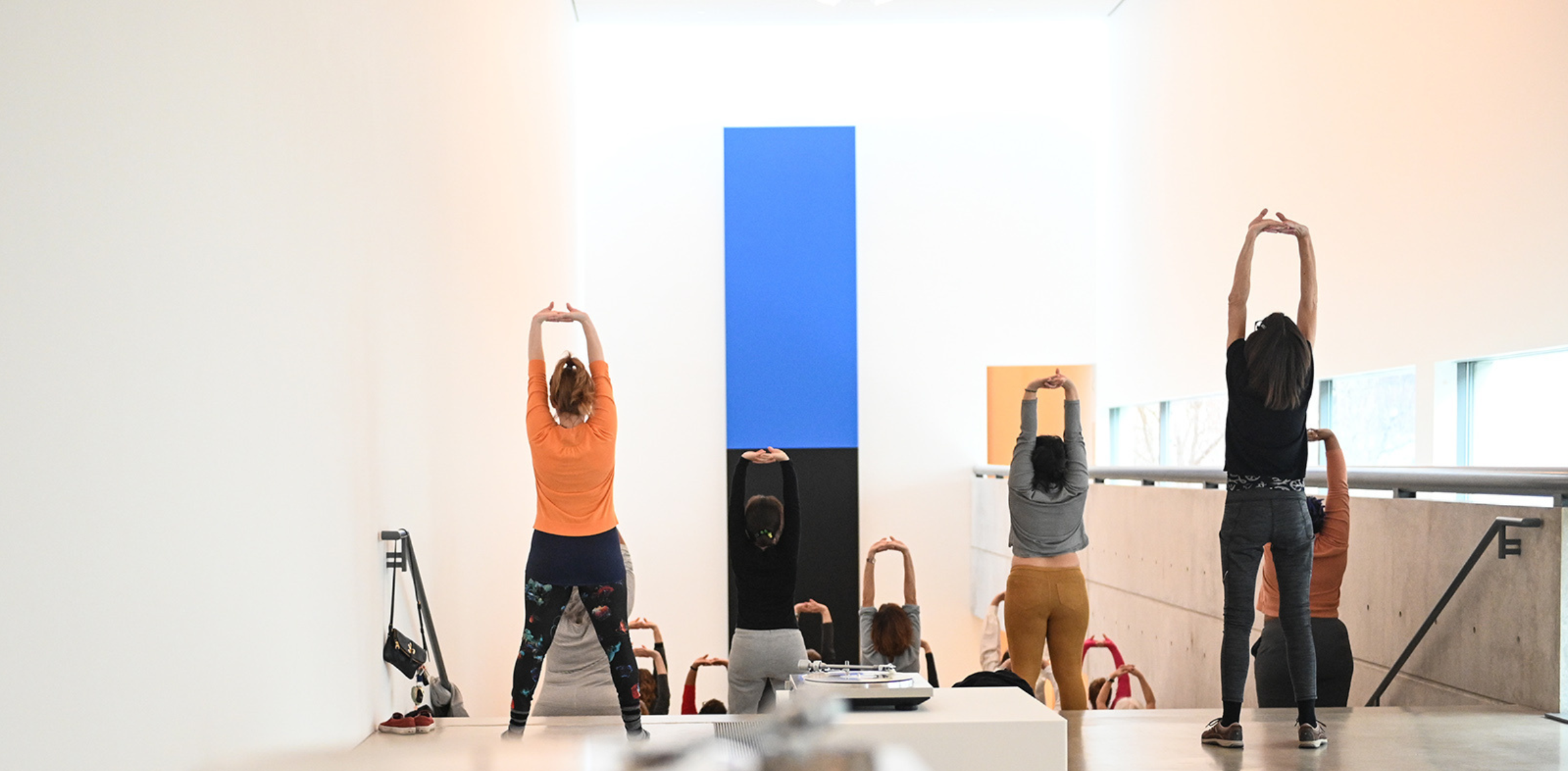 A group participates in a stretching session in a brightly lit gallery, framed by minimalist architecture and a bold blue and black artwork. Natural light fills the space, creating a calm and focused atmosphere.