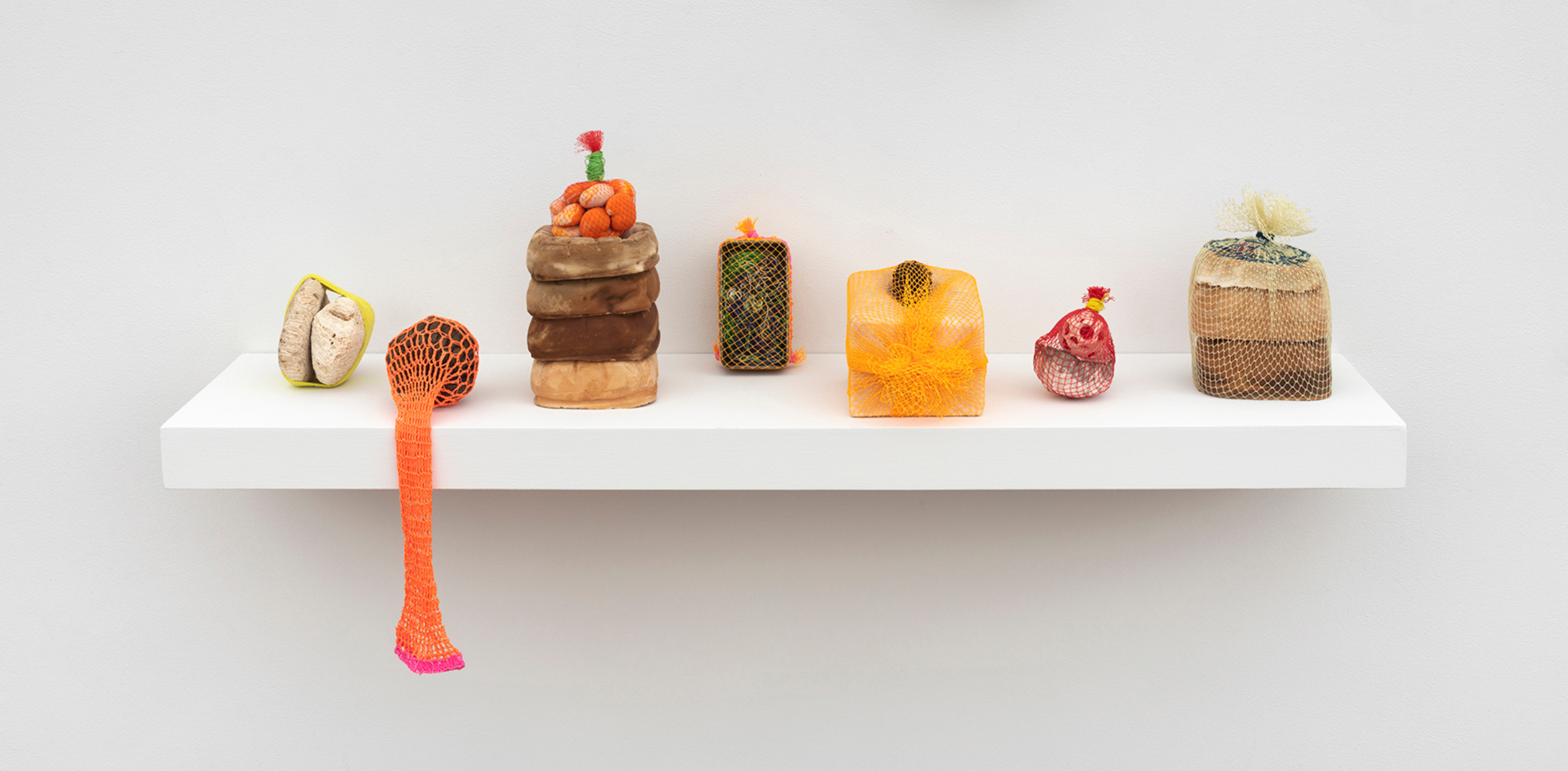 A row of small, colorful sculptures made from netted and textured materials, displayed on a white shelf against a plain wall.