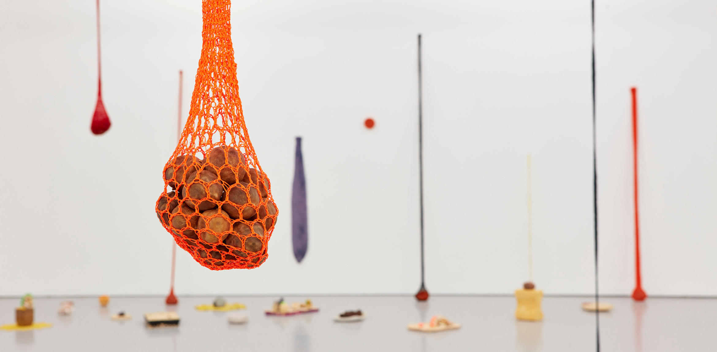 A close-up of an orange netted bag containing small objects, suspended in a gallery space with colorful hanging elements and sculptures blurred in the background.