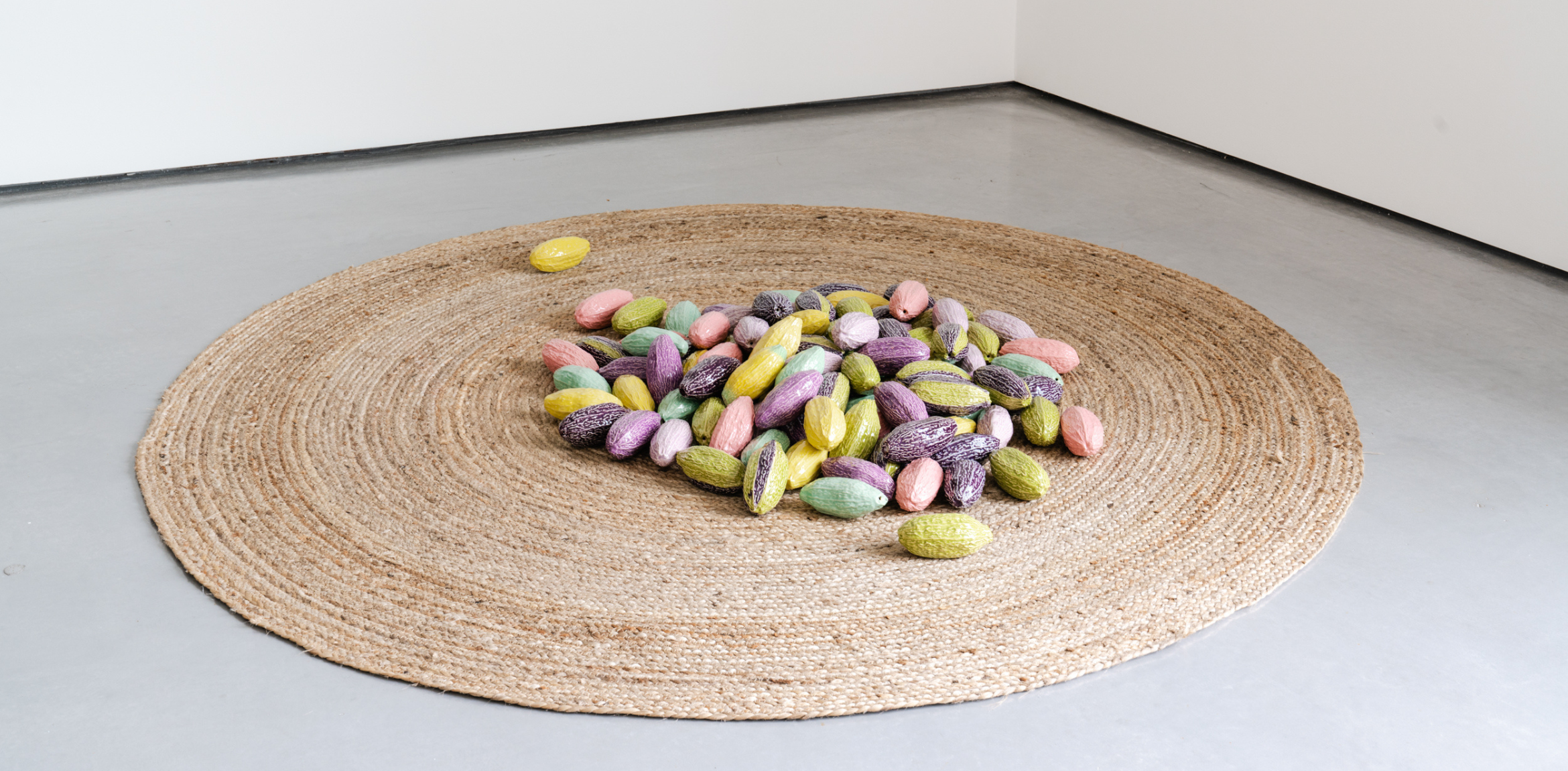 A pile of pastel-colored, textured sculptures resembling cocoa pods sits on a woven circular rug in a gallery space with a plain white wall and concrete floor. One pod is slightly separated from the group.