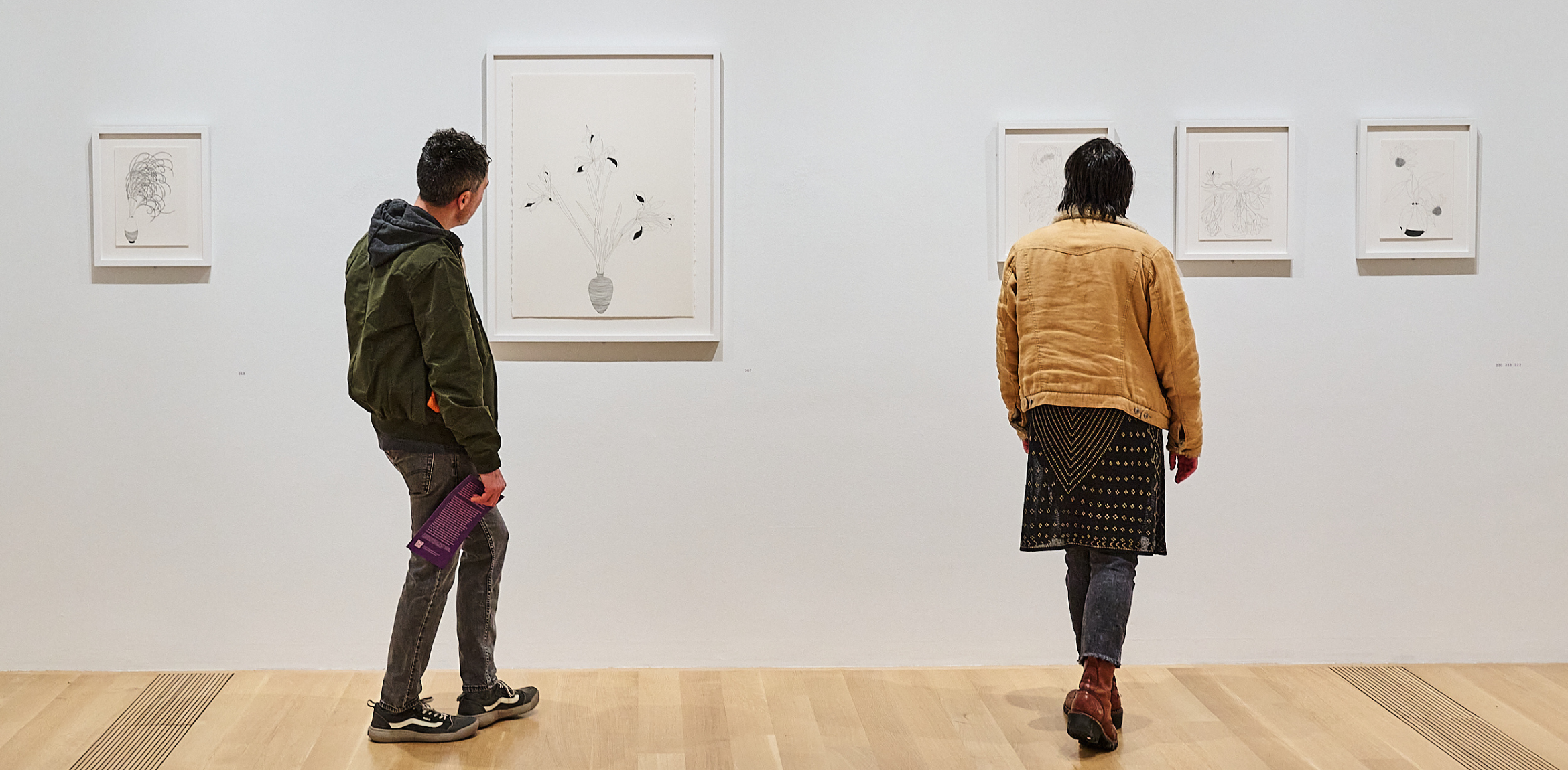 Two visitors view framed black-and-white drawings on a gallery wall.