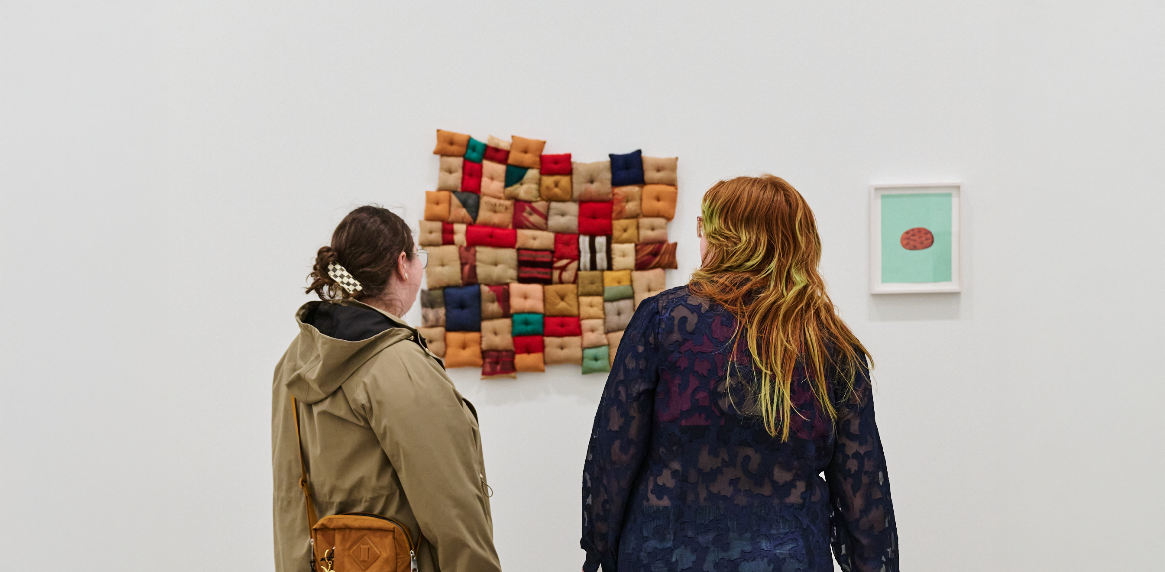 Two visitors view a wall-mounted textile artwork made of colorful fabric squares in a gallery.