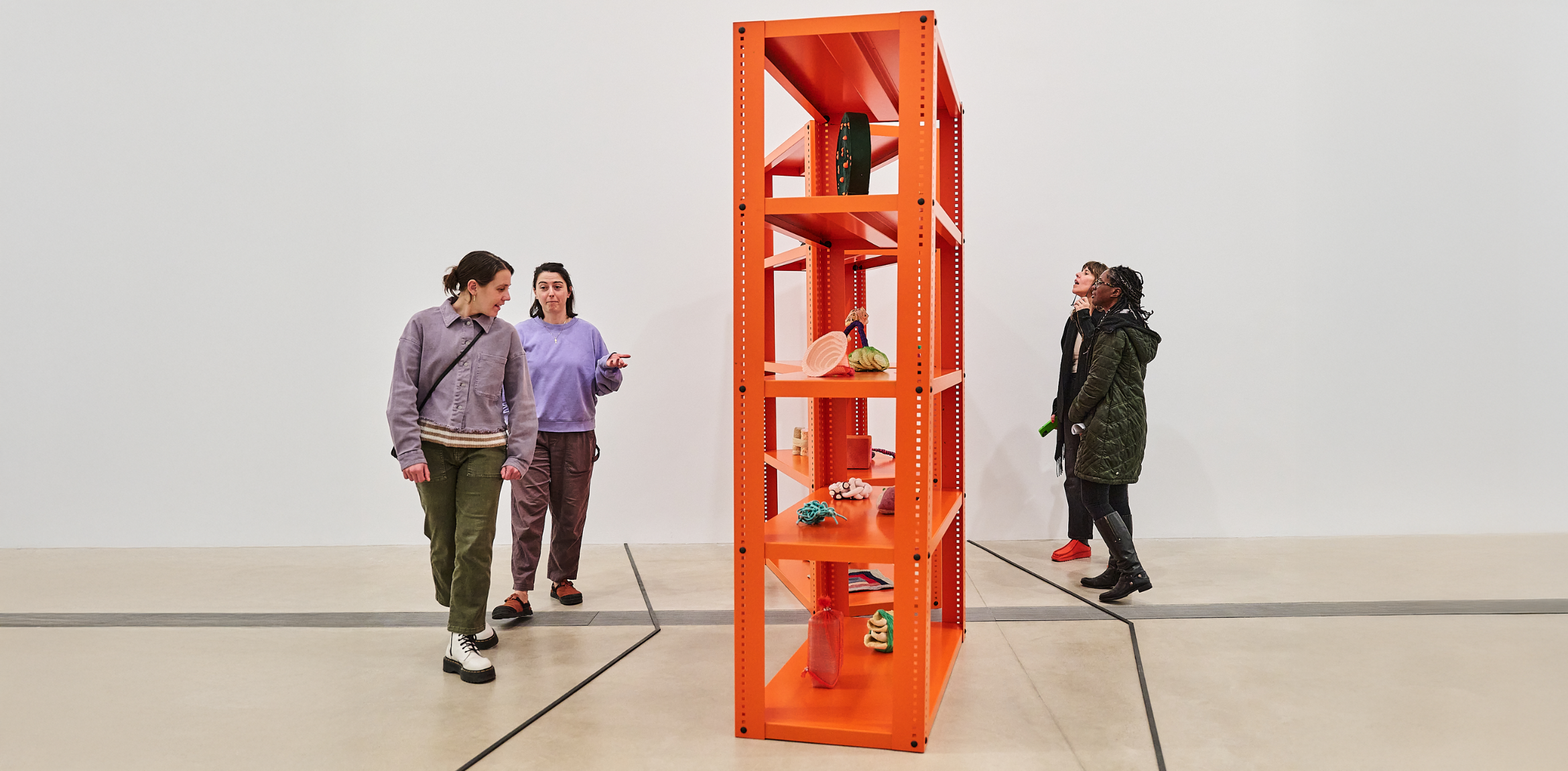 Visitors observe an orange shelving unit displaying small sculptural objects in a gallery space.