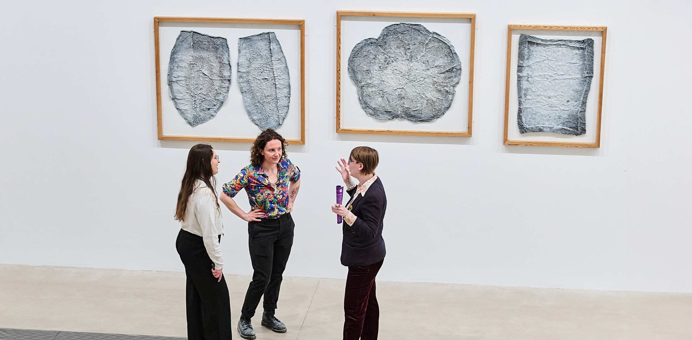 Three people stand in conversation in front of framed abstract artworks on a white gallery wall.