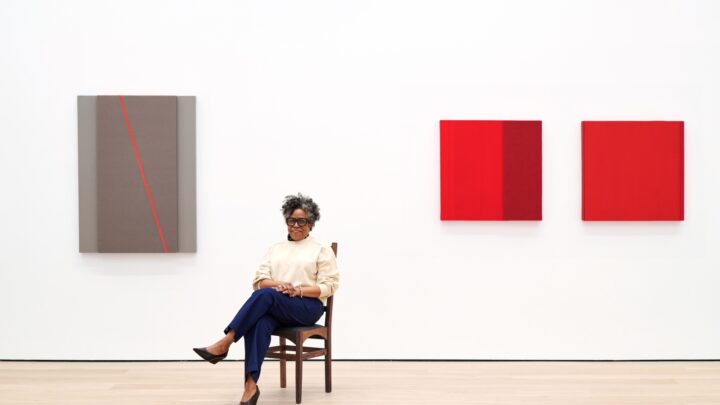 Person seated in front of three abstract wall-mounted artworks in red and gray tones, with light wood flooring and a white wall.