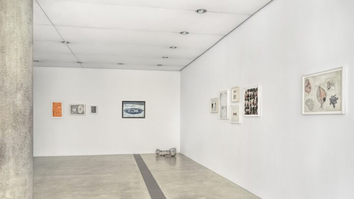 Gallery view with framed artworks arranged in a line on two adjacent white walls, with a small sculptural object placed on the floor near the corner.
