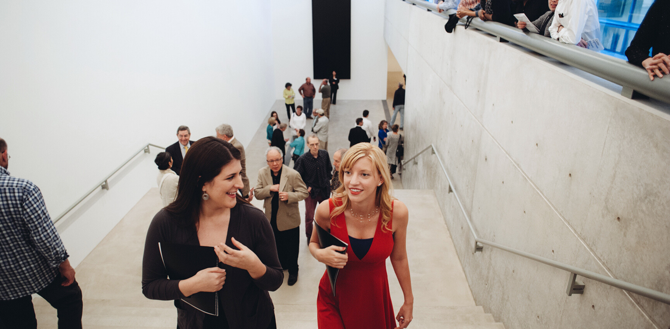 A visitor in with brown hair in a black dress and one with blonde hair in a red dress walking up the stairs at a Pulitzer opening.