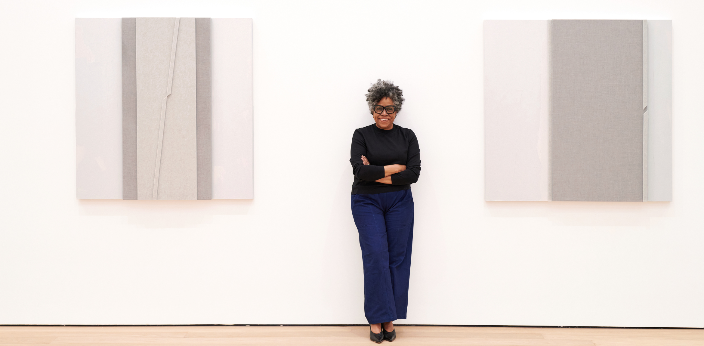 Artist Jennie C. Jones smiling and standing with her arms cross with her work hanging from the walls.