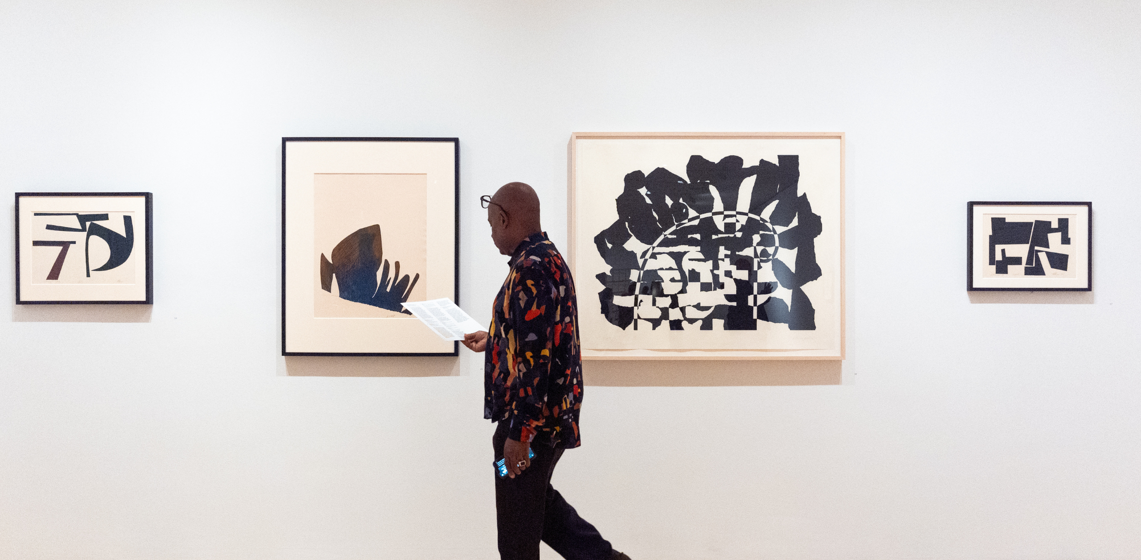 Man walking past black and white abstract artworks on a white gallery wall. He is looking down at a gallery card.