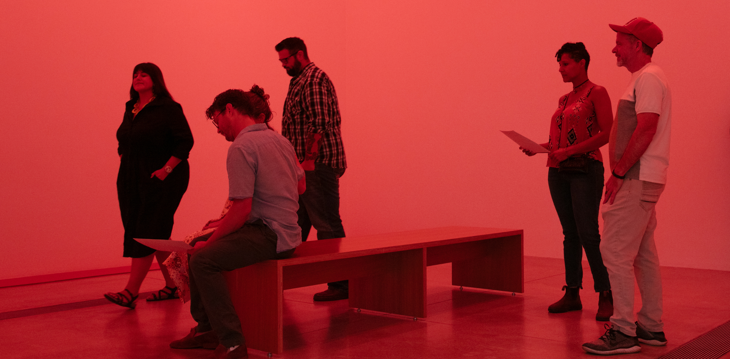 Visitors standing or sitting on a wooden bench in a red-lit room